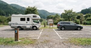 日本の山あいにあるRVパークの駐車区画に、カムロード架装のキャンピングカーと乗用車が真横から見た角度で並んで停まっている昼間の様子