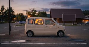 夕方の道の駅の駐車場に側面から見て停まる軽自動車、車内にほんのり灯りが灯っている様子