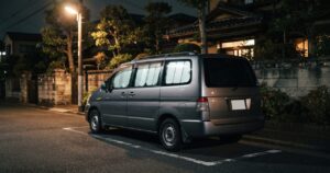 夜の住宅街の道路脇、街灯に照らされ目隠しシェードを付けたミニバンが後方斜めから見える様子