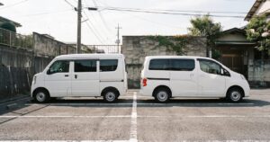 白い軽自動車の商用バンと一回り大きい白い商用バンが、住宅街の駐車場に真横向きで並んで停まっている様子