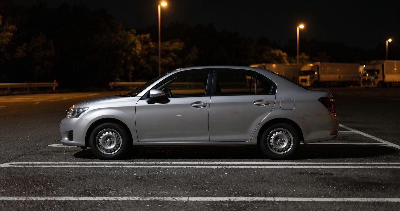 夜の高速道路サービスエリアの駐車場に側面から停まる乗用車、奥に大型トラックエリアの明かりが見える