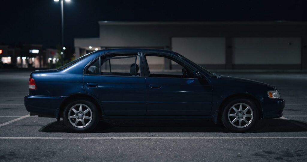 夜の駐車場に側面から停まる紺色の乗用車、後部座席の窓を開けて網目のウィンドウネットを取り付けている様子