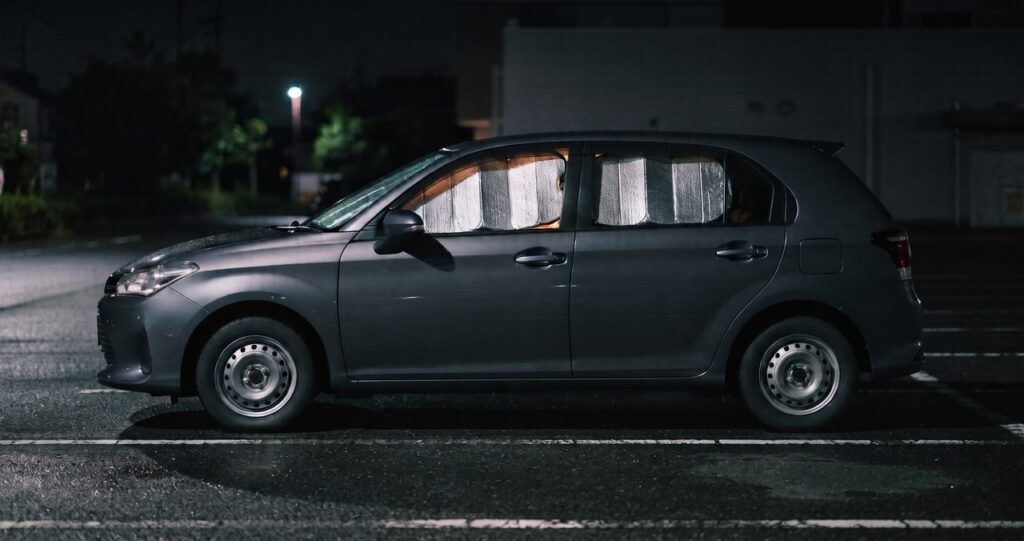 夜の駐車場に側面から停まる乗用車、フロントガラスと側面の窓に銀色の断熱シェードが取り付けられ車内からほのかな光が漏れている様子