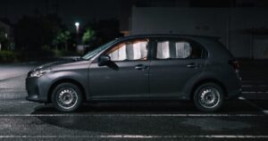 夜の駐車場に側面から停まる乗用車、フロントガラスと側面の窓に銀色の断熱シェードが取り付けられ車内からほのかな光が漏れている様子