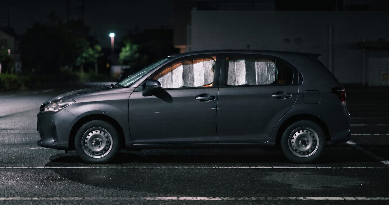 夜の駐車場に側面から停まる乗用車、フロントガラスと側面の窓に銀色の断熱シェードが取り付けられ車内からほのかな光が漏れている様子