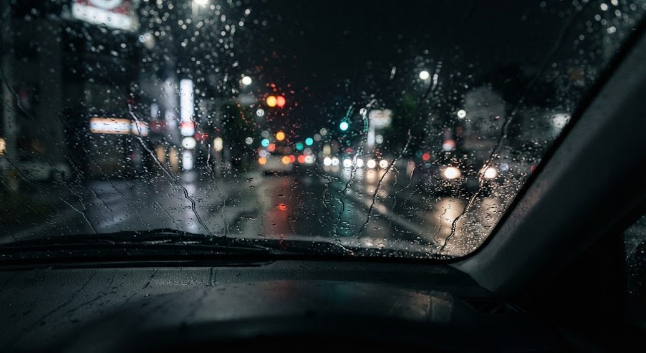雨の降る夜、車内から見たフロントガラスに雨粒が伝い落ち、外は暗く遠くの街灯や車のライトの光がにじんで見える様子