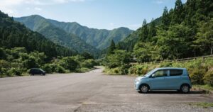 山あいの静かな道の駅の駐車場、手前に薄い水色の軽自動車が1台側面向きに停まり、奥にもう1台が小さく見える様子