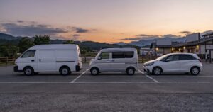 薄暮の駐車場に、真横から見た角度で停まる複数の車中泊仕様の車、道の駅やRVパークをイメージさせる開けた駐車スペース